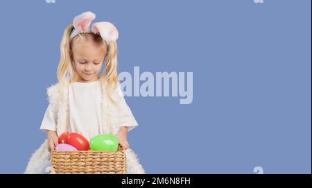 Hübsches, süßes kleines Mädchen mit Häschenohren. Das Kind hält sich in den Händen und schaut in den Osterkorb mit Eiern auf blauem Hintergrund. Speicherplatz kopieren. Stockfoto