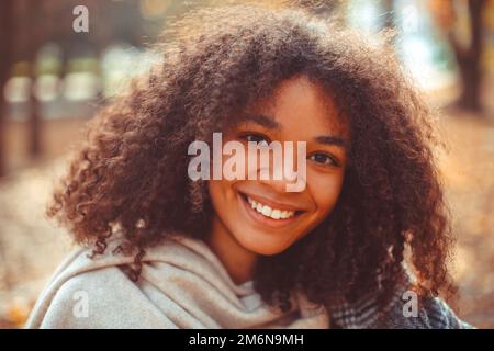 Süßes Herbstporträt einer jungen, lächelnden, glücklichen afroamerikanischen Frau mit lockigem Haar, die im Herbst im Park spazieren geht Stockfoto