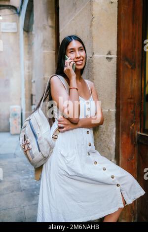 Optimistische hispanische Reisende in weißem Kleid mit Rucksack, der mit einem Lächeln wegschaut und sich mit einem Smartphone unterhält, während er sich an die Wand eines alten Gebäudes in der Nähe der Holztür der Straße von Barcelona, Spanien, lehnt Stockfoto