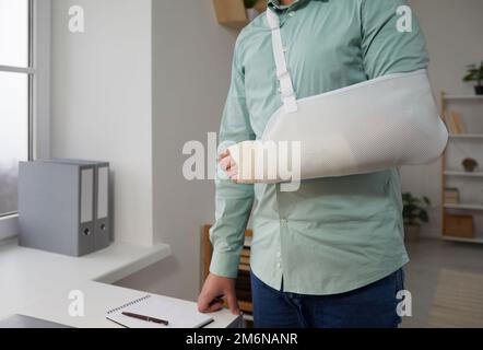 Zugeschnittenes Bild eines Mannes mit Verbandshand, der neben dem Schreibtisch am Arbeitsplatz im Büro steht. Stockfoto