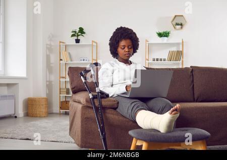 Eine attraktive afroamerikanische Frau mit gebrochenem Bein im Gips sitzt auf dem Sofa im Wohnzimmer mit einem Laptop in der Hand. Stockfoto