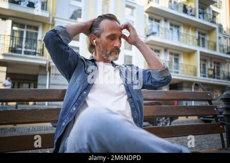 Ein gutaussehender Mann mittleren Alters sitzt auf der Bank und hört Musik mit kabellosen Kopfhörern, wobei die Hände hochgehoben und sein langes Haar geflickt sind Stockfoto