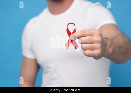 Mann mit rotem Schleifenband AIDS Wahrnehmungssymbol in der Hand trägt ein weißes T-Shirt isoliert auf blauem Hintergrund. Moderne Medizin und Stockfoto