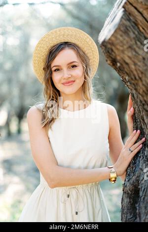 Ein Mädchen mit Strohhut steht neben einem Baum in einem Olivenhain. Porträt Stockfoto