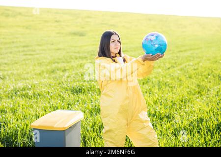 Eine Frau in einem gelben Schutzanzug steht mitten auf einem grünen Feld und hält einen Globus in den Händen, neben einer Mülltonne. Planet Care, speichern Sie die Stockfoto