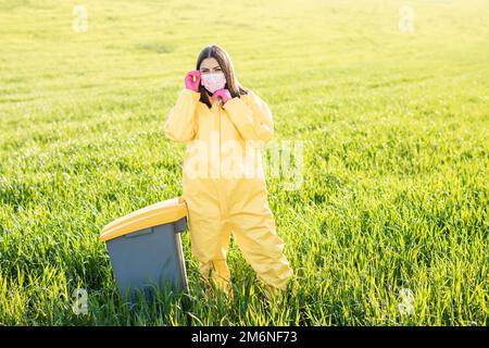 Eine Frau in einem gelben Schutzanzug steht in einer Schutzmaske in der Mitte eines grünen Feldes und trägt rosa Handschuhe. Stockfoto