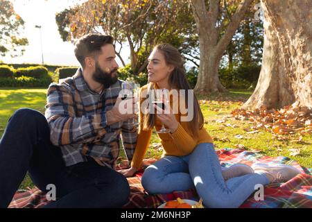 Glückliches kaukasisches Paar, das im sonnigen Herbstgarten auf dem Teppich sitzt und Wein trinkt Stockfoto