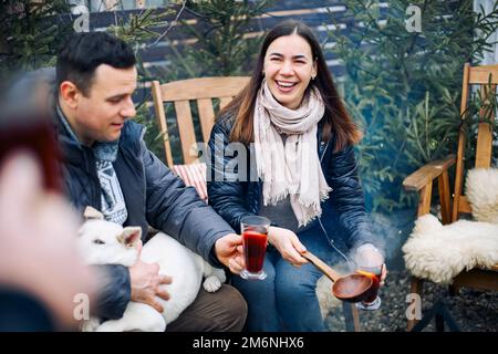 Winterparty mit Lagerfeuer. Fröhliches junges Paar mit Hund, das Glühwein in Gesellschaft von Freunden trinkt Stockfoto