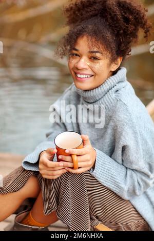 Junge, fröhliche afroamerikanische Frau mit einer Tasse heißem Tee, die sich im Herbst in der Nähe des Teichs entspannt, gemischte Rasse, weiblich in warmem Swe Stockfoto