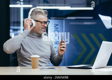 Leitender, grauhaariger Geschäftsmann, der in einem modernen Büro mit einem Laptop arbeitet, ein erwachsener Mann mit Kopfhörern, der Musik hört und Podcasts von Hörbüchern über die App auf dem Telefon anhört. Stockfoto
