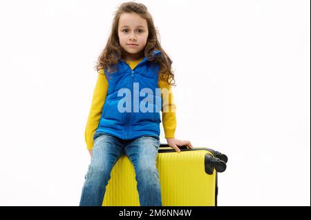 Ein bezauberndes Mädchen in blauer Jacke und Jeans, sitzt auf einem gelben Koffer, schaut in die Kamera, isoliert auf weißem Hintergrund. Stockfoto