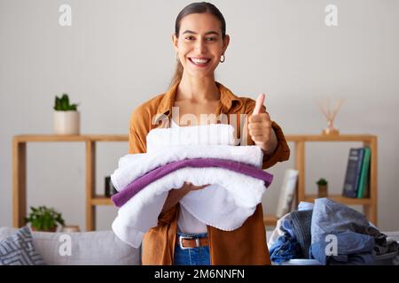 Daumen hoch, Wäsche und Porträt eines weiblichen Zimmermädchens, das Kleider im Wohnzimmer in einem modernen Haus faltete. Glücklich, lächeln und weibliche Putzfrau oder Hausfrau Stockfoto