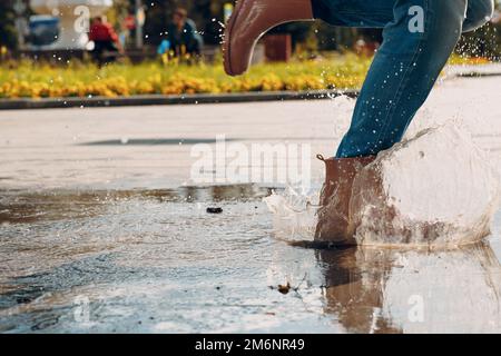 Frau mit Regengummistiefeln, die beim Herbstregen mit Wasserspritzern und Tropfen in die Pfütze laufen und springen Stockfoto