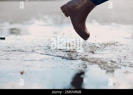 Frau mit Regengummistiefeln, die beim Herbstregen mit Wasserspritzern und Tropfen in die Pfütze laufen und springen Stockfoto