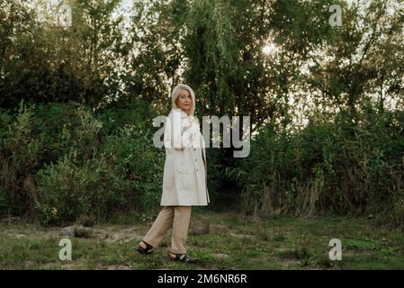 Eine schöne Erwachsene Frau mit blondem Haar und einem leichten Regenmantel lächelt Stockfoto