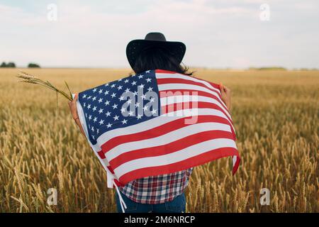 Farmerin trägt Cowboyhut, kariertes Hemd und Jeans mit amerikanischer Flagge auf dem Weizenfeld Stockfoto