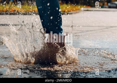 Frau mit Regengummistiefeln, die beim Herbstregen mit Wasserspritzern und Tropfen in die Pfütze laufen und springen Stockfoto