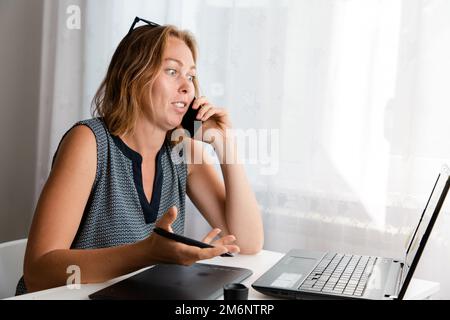 Remote-Arbeit. Eine Frau arbeitet zu Hause auf einem grafischen Tablet und spricht überraschend am Telefon. Das Konzept von Quarantäne, Freiberufung und Selbstisolation. Stockfoto