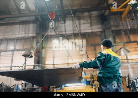 Arbeiter heben Metallblech mit Krankettenzug mit Fernbedienung und Haken in der industriellen Fabrik. Stockfoto