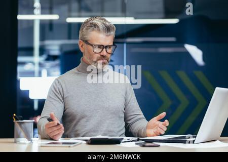 Unzufriedener, grauhaariger Chef im Büro, leitender Mann enttäuscht von dem erreichten Ergebnis, reifer Geschäftsmann verärgert und traurig, mit Dokumenten zu arbeiten und Laptop im Gebäude zu verwenden. Stockfoto