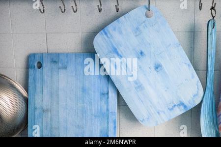 Nahaufnahme von zwei Holzschneidebrettern, die an einer gefliesten Wand an Metallhaken hängen, blau getönt Stockfoto