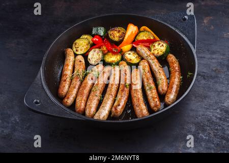 Traditionelle gebratene deutsche Schweinebratwurst mit Barbecue-Gemüse, serviert als Nahaufnahme in einer gusseisernen Pfanne auf rustikalem Schwarz Stockfoto