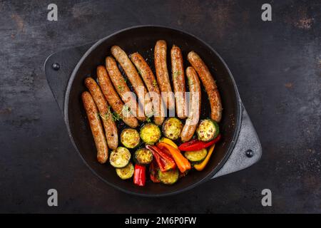 Traditionelle gebratene deutsche Schweinebratwurst mit Barbecue-Gemüse, serviert als Draufsicht in gusseiserner Pfanne auf rustikalem Schwarz Stockfoto