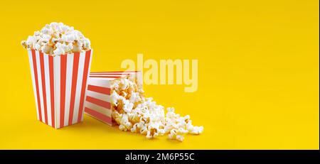 Zwei rot-weiß gestreifte Eimer mit leckerem Käsepopcorn, isoliert auf gelbem Hintergrund. Schachtel mit Streuung von Popcornkörnern. Filme, Kino A. Stockfoto