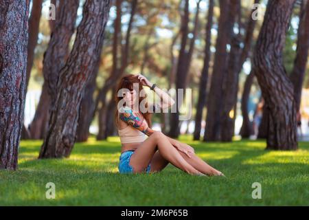 Eine weiße, wunderschöne, junge, gebräunte Frau mit Tattoos sitzt auf dem Rasen in einem Parkhain. Sommerurlaub im Freien. Stockfoto