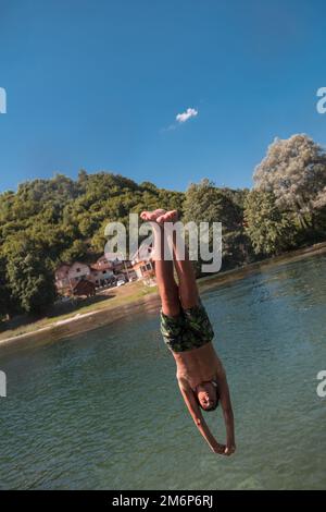 Junge Teenager springen fliegen und tauchen im Fluss. Klarer blauer Himmel und Bäume in der Ferne als natürlicher Hintergrund. Stockfoto