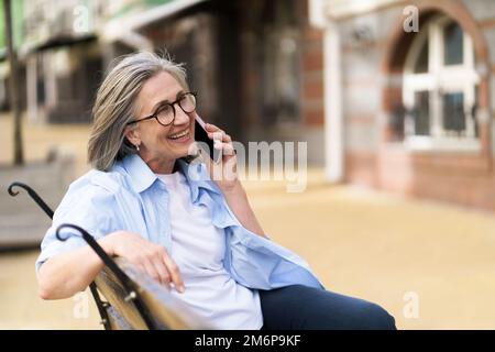 Reife grauhaarige Frau, die am Telefon lächelt, auf der Bank sitzt und die Sommerzeit auf den Straßen des alten europäischen Schleppnetzes genießt Stockfoto