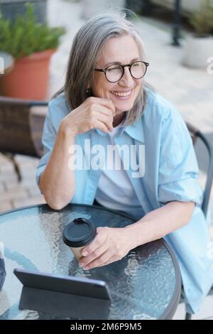 Grauhaarige, reife, europäische lächelnde Frau trinkt Kaffee mit einem digitalen Tablet, das am Tisch des Straßencafés sitzt. Reifes Silberhaar Stockfoto