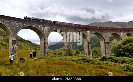 Menschen beobachten und winken der Jacobite Dampfeisenbahn, die das Glenfinnan Viadukt im schottischen Hochland durchquert. Stockfoto