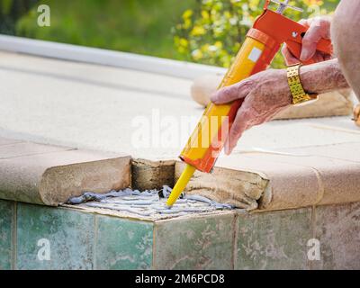 Mann, der Zementkleber mit einer Fugenpistole aufträgt. Reparatur von gebrochenen Fliesen aus Betonsteinen im Schwimmbad. Stockfoto