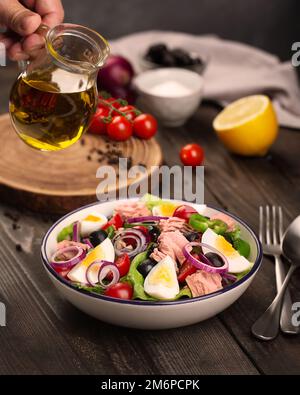 Nicoise-Salat mit Thunfisch, Eiern, Oliven und Salatblättern auf einem Holztisch. Die Hand eines Mannes hält eine kleine Kanne Olivenöl Stockfoto