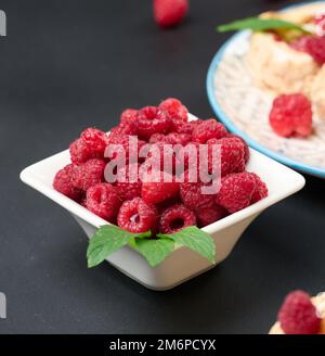 Reife rote Himbeeren in einer weißen Keramikschale auf einem schwarzen Tisch Stockfoto