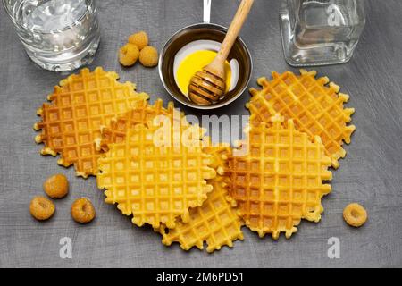 Wiener Waffeln und gelbe Himbeeren auf dem Tisch. Metallschüssel mit Honig und Honigstab aus Holz. Glas und Flasche mit Wasser. Flach verlegt. Grauer Hintergrund Stockfoto