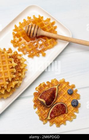 Feigenscheiben und Beeren auf einer Waffel. Waffeln mit Honig und Honigstäbchen auf einem weißen Teller. Flach verlegt. Weißer Hintergrund. Stockfoto