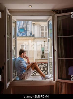 Ein junger Mann sitzt in einem Fenster mit Blick auf Paris. Männer genießen die Sonne im Fenster des Hotelzimmers Stockfoto