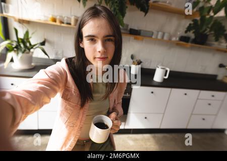 Porträt einer nicht-binären Transfrau, die eine Kaffeetasse in der Hand hält und die Kamera in der Küche zu Hause anschaut Stockfoto