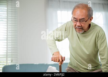 Asiatischer alter Mann mit Brille, der tippt, um vom Sofa mit Gehstock aufzustehen Stockfoto