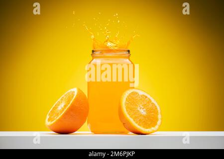 Frischer Orangensaft in einem Glasgefäß mit Spritzern und Orangenscheiben auf einem Regal mit gelbem Hintergrund. Stockfoto