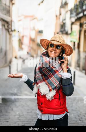 Eine Frau mittleren Alters mit Hut und gelber Sonnenbrille, die telefoniert und auf der Straße lächelt. Konzept von Glück und Wohlbefinden. Stockfoto