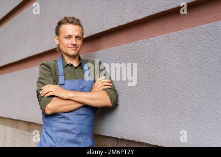 Ein attraktiver Angestellter, der mit zusammengefalteten Armen und einer Schürze draußen steht, macht eine Pause, lehnt sich an eine Wand und genießt die frische Luft nach dem Lon Stockfoto