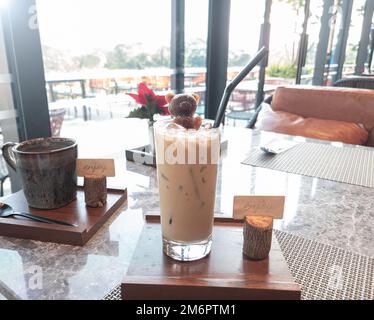 Ein Glas Latte-Kaffee mit einem Bärensirup-Eis auf Holzteller im Café und Blick auf den Außenbereich. Stockfoto
