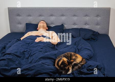 Seitenansicht eines gutaussehenden jungen Mannes, der nachts mit einer Katze auf einem Bett schläft. Junggesellenschlafzimmer. Tiefer Schlaf. Stockfoto