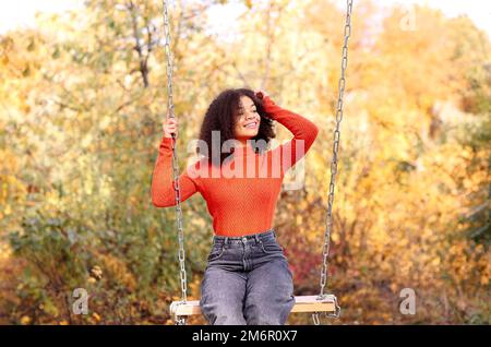 Junge, fröhliche, sorgenfreie afroamerikanische Frau mit lockigem Haar in orangefarbenem, warmem Pullover, die auf der Schaukel sitzt Stockfoto