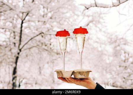 Glückliche ältere Frau in warmen Kleidern, die zwei Gläser Champagner hielt, während sie im verschneiten Innenhof stand Stockfoto