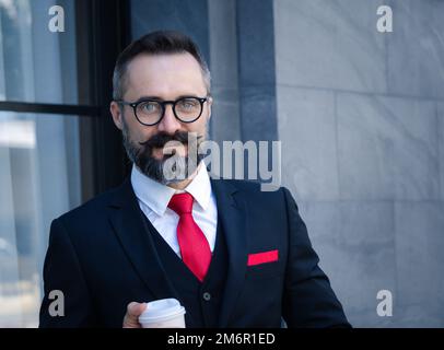 Porträt eines bärtigen Hipster-Geschäftsmannes im Anzug, der in einem Geschäftsviertel glücklich Kaffee trinkt. Stockfoto