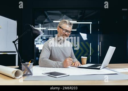 Erfolgreicher leitender Architekt bei der Arbeit im Büro, reifer, grauhaariger Mann, der Entwürfe zum Layout zeichnet und Pläne lächelt und mit der Arbeit zufrieden ist. Stockfoto